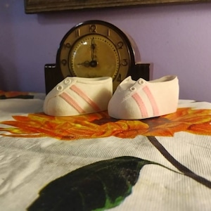 May include: Two white ceramic shoes with pink stripes, sitting on a white tablecloth with a floral pattern.