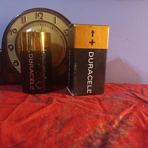 May include: A vintage Duracell battery and a battery-shaped bottle with a gold cap are displayed in front of a clock. The battery box is black and gold with the Duracell logo and a plus sign.