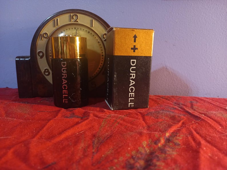 May include: A vintage Duracell battery and a battery-shaped bottle with a gold cap are displayed in front of a clock. The battery box is black and gold with the Duracell logo and a plus sign.