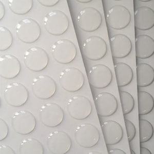 Może przedstawiać: Arkusz przezroczystych kropek samoprzylepnych, ułożonych w rzędy na białym tle.