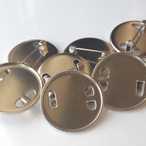 Könnte beinhalten: Eine Ansammlung glänzender, runder Metall-Anstecknadel-Rückseiten mit Sicherheitsnadelverschlüssen. Die Rückseiten haben zwei kleine Löcher und einen rechteckigen Schlitz zum Befestigen an einem Abzeichen. Die Nadeln liegen auf einer weißen Oberfläche.