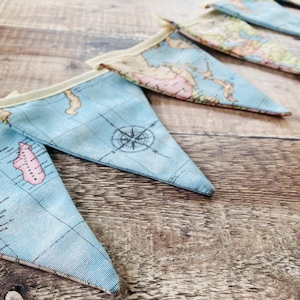 May include: A fabric bunting banner featuring triangular pennants with a vintage world map print. The flags are light blue with tan and pink landmass details, black grid lines, and a compass rose design, all attached to a cream-coloured fabric header.