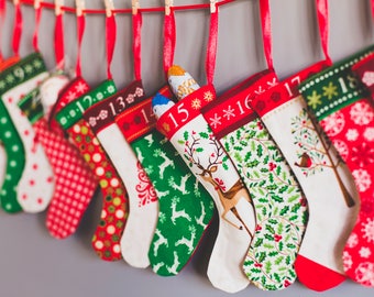 May include: A row of hanging Christmas stockings in red, green, and white, each featuring a unique holiday pattern and a numbered cuff from 9 to 19. The stockings are suspended from a red ribbon with small wooden clips, displaying festive motifs.