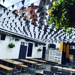 May include: A black and white striped awning with the text "The Old George" hangs over a patio with wooden tables and chairs. The patio is decorated with black and white triangle flags.