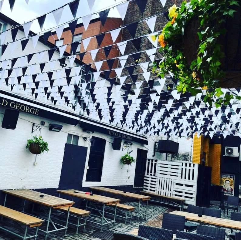 May include: A black and white striped awning with the text "The Old George" hangs over a patio with wooden tables and chairs. The patio is decorated with black and white triangle flags.