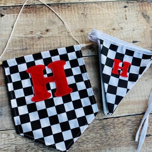 May include: Two checkered flags with a red letter "H" on a wooden background. The flags are black and white, with a pennant shape. One flag is larger and attached to a wooden dowel with string, and the other is smaller with white ties.