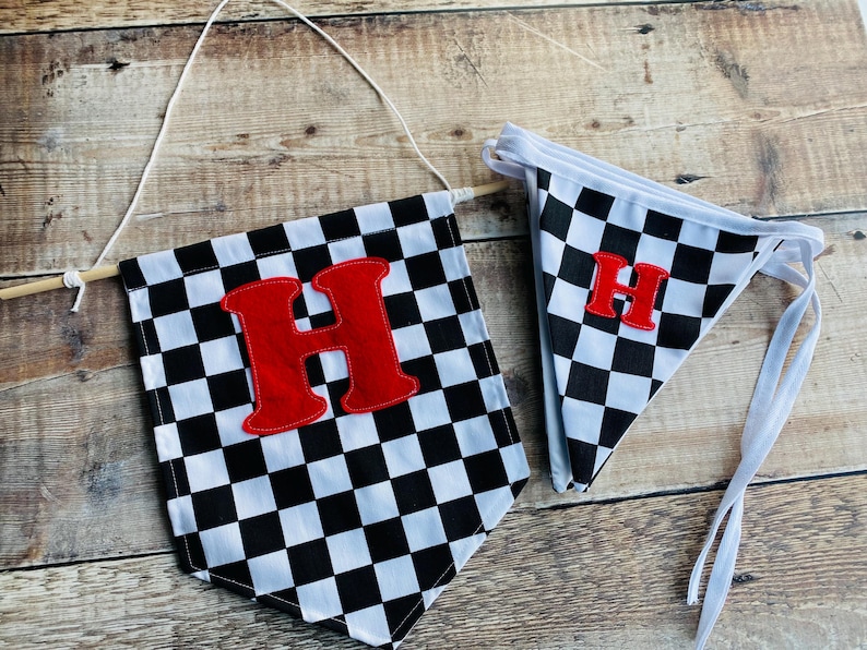 May include: Two checkered flags with a red letter "H" on a wooden background. The flags are black and white, with a pennant shape. One flag is larger and attached to a wooden dowel with string, and the other is smaller with white ties.