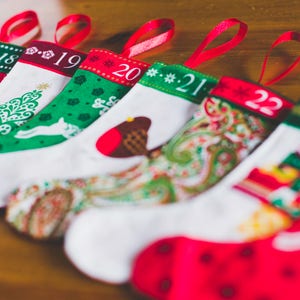 May include: A row of Christmas stockings with red ribbon loops. Each stocking is white with a colorful design and a number from 17 to 23. Designs include snowflakes, trees, and a bird.