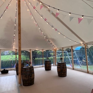 May include: A white tent with string lights and pink and white triangle flags hanging from the ceiling. There are four wooden barrels in the center of the tent.