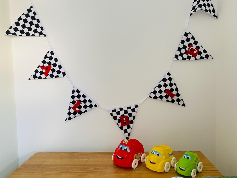 May include: A festive banner of black and white checkered flags with the letter "1" in red on each flag. Three toy cars in red, yellow, and green are below the banner, creating a racing theme. The banner and toys are on a wooden surface.