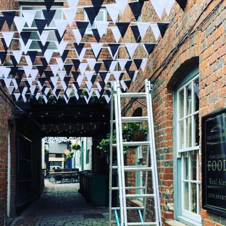May include: A brick alleyway decorated with black and white triangle bunting. A ladder is leaning against a brick wall with a sign that reads "The Old Garden, Great Quality Food, Real Ales, Live Sport".