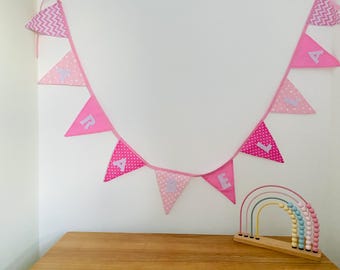 May include: A pink fabric banner with the letters spelling out a name, hanging against a white wall. The banner has triangle flags with polka dots and chevron patterns. A wooden rainbow abacus sits below.