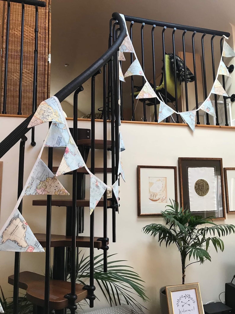 May include: A white and blue map-themed banner hangs on a black metal staircase railing. The banner is made of fabric and has a scalloped edge. The banner is decorated with a map of the world. The banner is hanging on a staircase railing in a home.