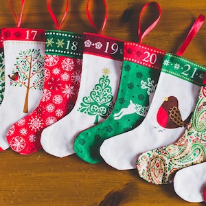 May include: A collection of Christmas stockings with red ribbon loops, each featuring a unique design and number from 15 to 22. Designs include snowflakes, trees, and birds, with red, green, and white color schemes.