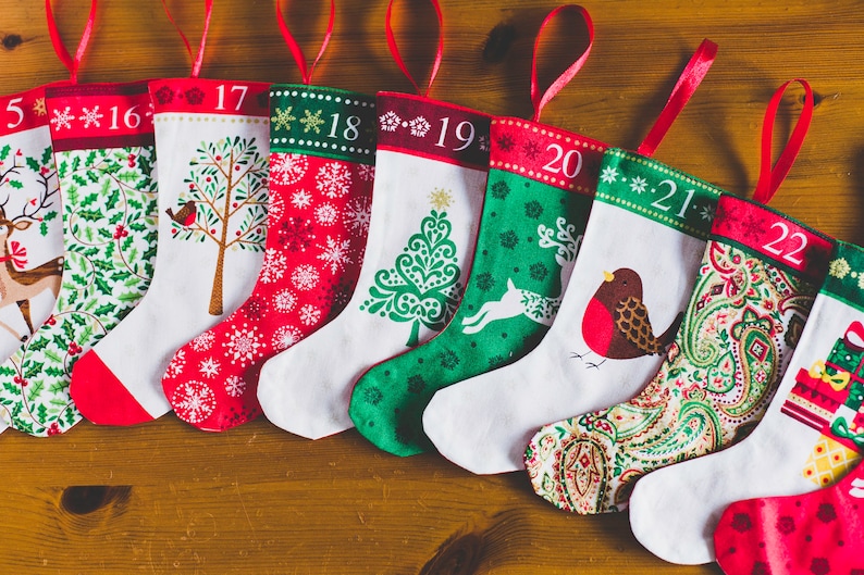 May include: A collection of Christmas stockings with red ribbon loops, each featuring a unique design and number from 15 to 22. Designs include snowflakes, trees, and birds, with red, green, and white color schemes.