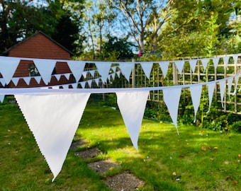 Op de afbeelding: Witte stoffen slinger met driehoekige vlaggen, gespannen over een groene tuin. De vlaggen zijn scherp, met een vage achtergrond van bomen, een houten schuur en een trellis. Perfect voor buitenfeesten.