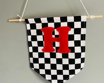 May include: A checkered flag banner with a large red letter "H" in the center. The banner is black and white, with a wooden dowel at the top and a string for hanging. The banner is likely for a nursery or child's room.