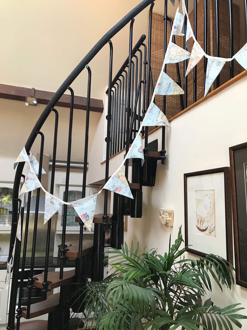 May include: A white and blue map-themed bunting hangs on a black metal spiral staircase. The bunting is strung between the railing and the wall. A green plant is in the foreground.