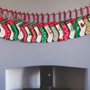 May include: A festive Christmas advent calendar featuring 24 small stockings, each with a unique pattern and number. The stockings are red, green, and white, and are suspended from a red string with wooden clothespins. The stockings are hung above a fireplace.