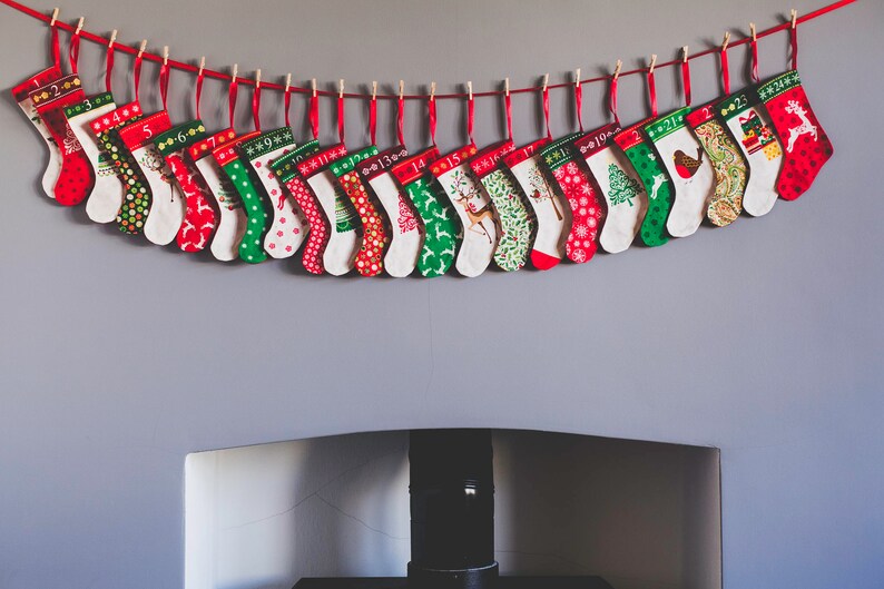 May include: A festive Christmas advent calendar featuring 24 small stockings, each with a unique pattern and number. The stockings are red, green, and white, and are suspended from a red string with wooden clothespins. The stockings are hung above a fireplace.