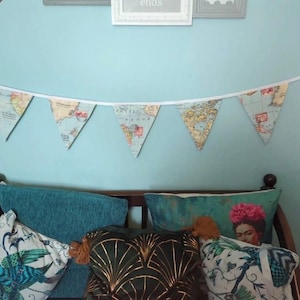May include: A teal blue couch with a variety of throw pillows. The pillows have a variety of patterns, including a green and gold geometric pattern, a floral pattern with a woman's face, and a bird pattern. A map-themed banner hangs above the couch.