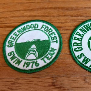 May include: Two circular embroidered patches. One features a shark graphic and the text "GREENWOOD FOREST SWIM TEAM 1976". The other patch has "GREENWOOD FOREST SWIM TEAM 1977" in a circle. Both have green and white colors.