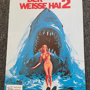 May include: Poster for "Der Weisse Hai 2" featuring a large blue shark with open jaws looming over a woman water skiing. The poster has the title in red and German text. The woman is wearing a yellow bikini.