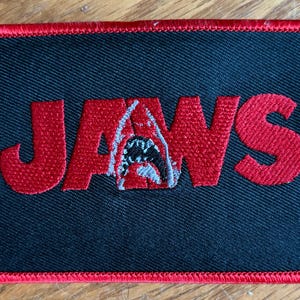 May include: A rectangular embroidered patch with a black background and a red border. The word JAWS is stitched in red, with the letter A replaced by a grey and white shark graphic featuring an open mouth with visible teeth and a dark interior.