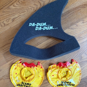 May include: A dark gray foam shark fin with the text "DA-DUM... DA-DUM..." in light green. Two yellow and red inflatable drink holders with the text "ALAMO DRAFTHOUSE" are also in the image.