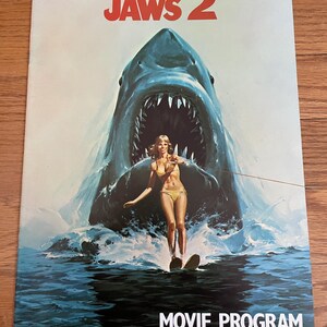 May include: A vintage movie program for "Jaws 2" featuring a woman water skiing in front of a large shark with an open mouth. The title "JAWS 2" is in red at the top, and "MOVIE PROGRAM" is at the bottom.