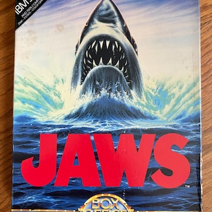 May include: A rectangular Jaws computer game box for IBM 3 1/2 inch disks. The cover features a large shark emerging from blue ocean water with white splashes. The title JAWS is printed in large red letters, with the text BOX OFFICE below it.