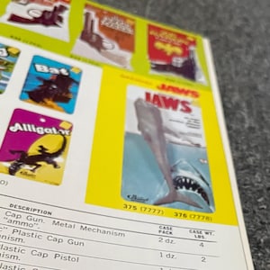 May include: A product display featuring toy packaging with the word "JAWS" prominently displayed. The packaging shows a shark illustration. Other product labels include "Bat" and "Alligator". The display also includes product descriptions and case pack information.