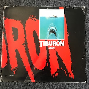 May include: A red and black poster with the words "Tiburon" and "Jaws" in white. The poster features a close-up of a great white shark's head and mouth.