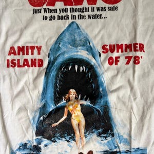 May include: A white t-shirt featuring a graphic of a large shark emerging from water behind a person on water skis. Text includes JAWS, Just When you thought it was safe to go back in the water, AMITY ISLAND, SUMMER OF 78, and MARTHA'S VINEYARD.