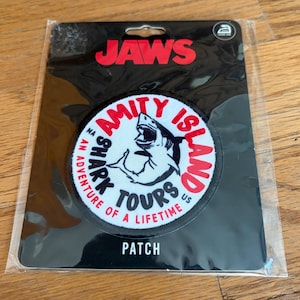 May include: A black and white patch featuring a shark graphic with the words "Amity Island Shark Tours" in red and white. The patch is packaged in a clear plastic sleeve with the word "PATCH" at the bottom.