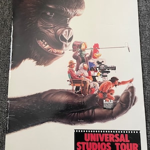 May include: A vintage Universal Studios Tour promotional poster featuring a large gorilla hand holding a group of movie characters, including a cowboy, an astronaut, a monster, and a cartoon rooster. The text on the poster reads "Universal Studios Tour, An MCA Company."