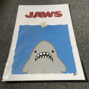 May include: A white poster with the word "JAWS" in red letters at the top. A cartoon shark with a toothy grin is in the center of the poster, looking up at a cartoon sandwich with a smiling face floating above the water.