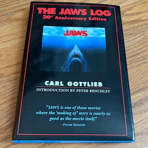 May include: A book titled "THE JAWS LOG 30th Anniversary Edition" featuring a shark movie poster. The author is Carl Gottlieb, with an introduction by Peter Benchley. A quote from Peter Biskind is also included on the cover.