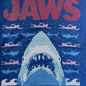 May include: A blue t-shirt with a knitted design of the word "JAWS" in red and a white shark fin with an open mouth. The shark is surrounded by blue and pink clouds.