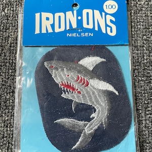 May include: A blue and white iron-on patch featuring a gray shark with red teeth. The patch is packaged in a clear plastic bag with a blue and white card that reads "IRON-ONS by NIELSEN 1.00".