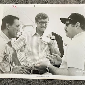May include: Black and white photo of three men. The man in the middle is wearing a plaid shirt and glasses. The man on the left is wearing a dark shirt. The man on the right is wearing a dark hat and a light shirt. The photo is captioned "Universal Pictures".