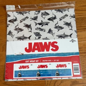 May include: A gift wrap kit featuring a shark-themed design. The wrapping paper has a pattern of gray sharks on a white background. The kit includes wrapping paper and gift tags with the word "JAWS" in red.
