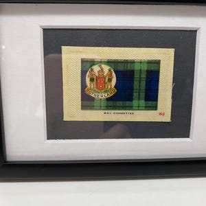 May include: A framed vintage cigarette card featuring a green, blue, and yellow tartan pattern with a crest that says "Sutherland" and the text "B.D.V. Cigarettes" and "63".