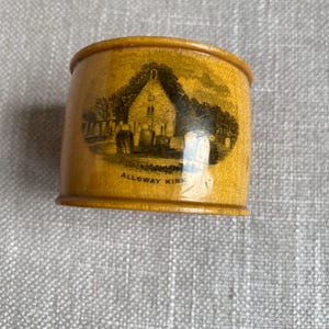 May include: A cylindrical, wooden napkin ring with a light brown finish. The ring features a black and white illustration of a church with the text "ALLOWAY KIRK" below the image. The design is a vintage style.