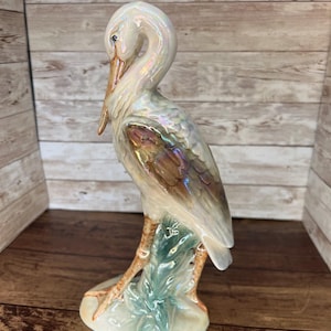 May include: A ceramic heron figurine with iridescent white, brown, and green hues. The bird stands on a base with green foliage. The heron has a long beak and is in a curved pose.