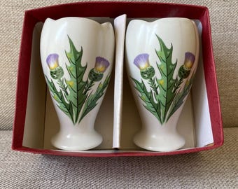 May include: Two white ceramic egg cups with a thistle design, presented in a red box. The thistle design features purple flowers and green leaves. The egg cups have a flared top and a pedestal base.