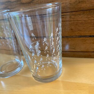 Op de afbeelding: Twee heldere drinkglazen met een witte afbeelding van een persoon die loopt. De glazen zijn cilindrisch en staan op een licht houten oppervlak. De tekst "KEES NVITING" is zichtbaar op de zijkant van een glas.