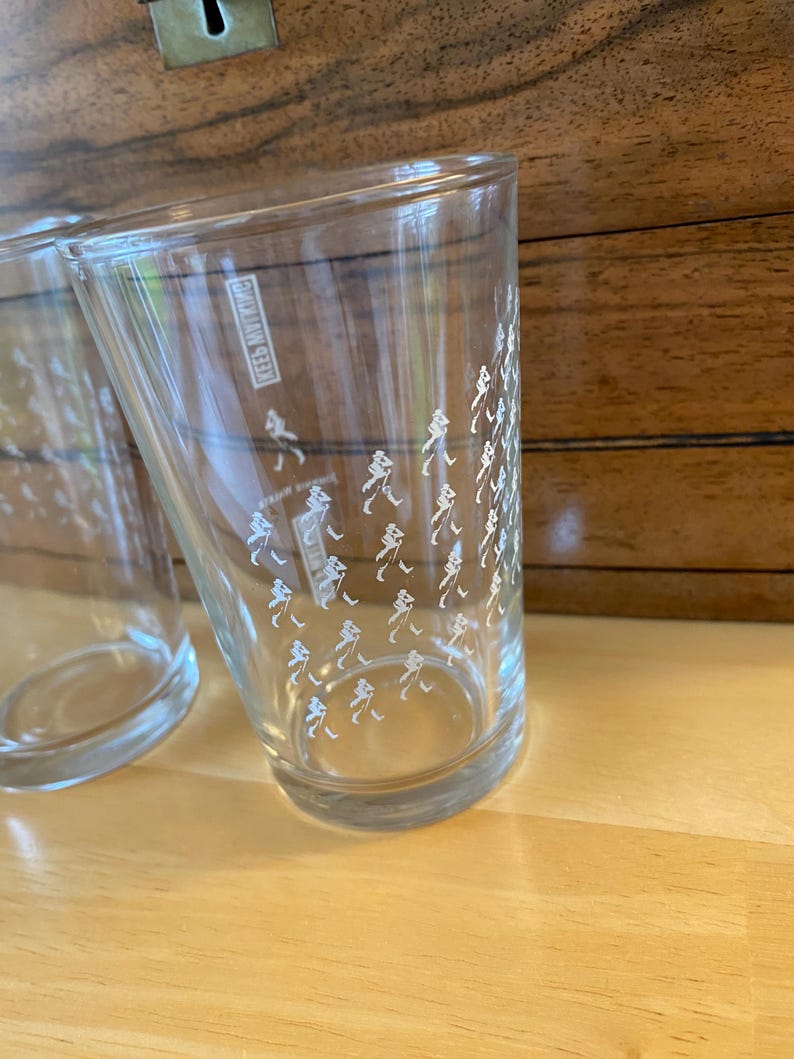 Op de afbeelding: Twee heldere drinkglazen met een witte afbeelding van een persoon die loopt. De glazen zijn cilindrisch en staan op een licht houten oppervlak. De tekst "KEES NVITING" is zichtbaar op de zijkant van een glas.