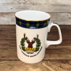 May include: White ceramic mug with a gold rim and a handle. The mug features a blue, green, and yellow tartan pattern at the top and a crest with a stag and the words "The Gordon Highlanders BYDAND".