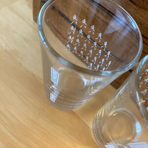 Op de afbeelding: Twee heldere glazen bekers met witte bedrukte ontwerpen. E&eacute;n beker heeft kleine figuren, de andere een gebouwontwerp. De glazen staan op een licht houten oppervlak.